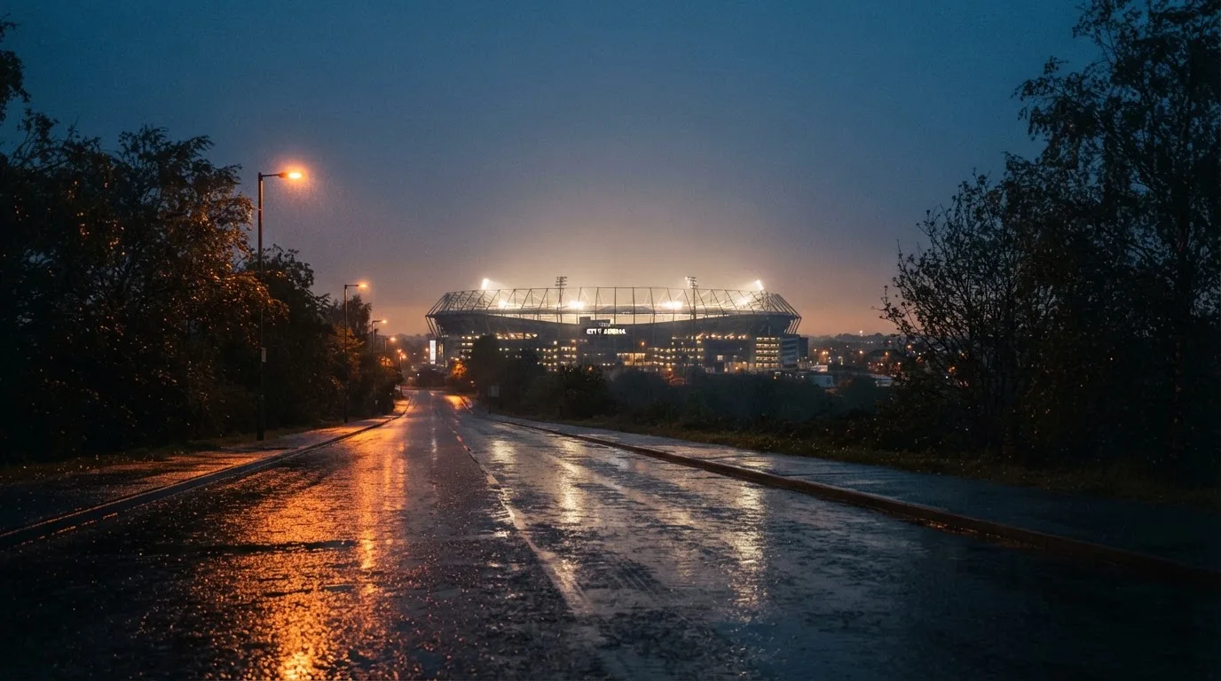 Ein Stadion unter Flutlicht aus großer Entfernung gesehen, davor eine regennasse Straße in der Dämmerung.