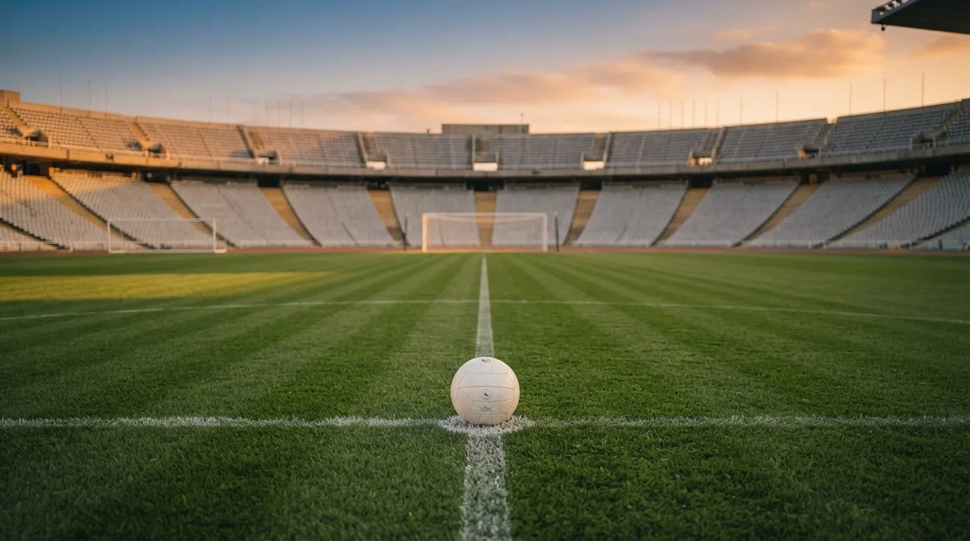 Ein Fußball liegt auf dem Anstoßpunkt eines leeren Rasenplatzes, im Hintergrund eine menschenleere Tribüne im weichen Abendlicht.