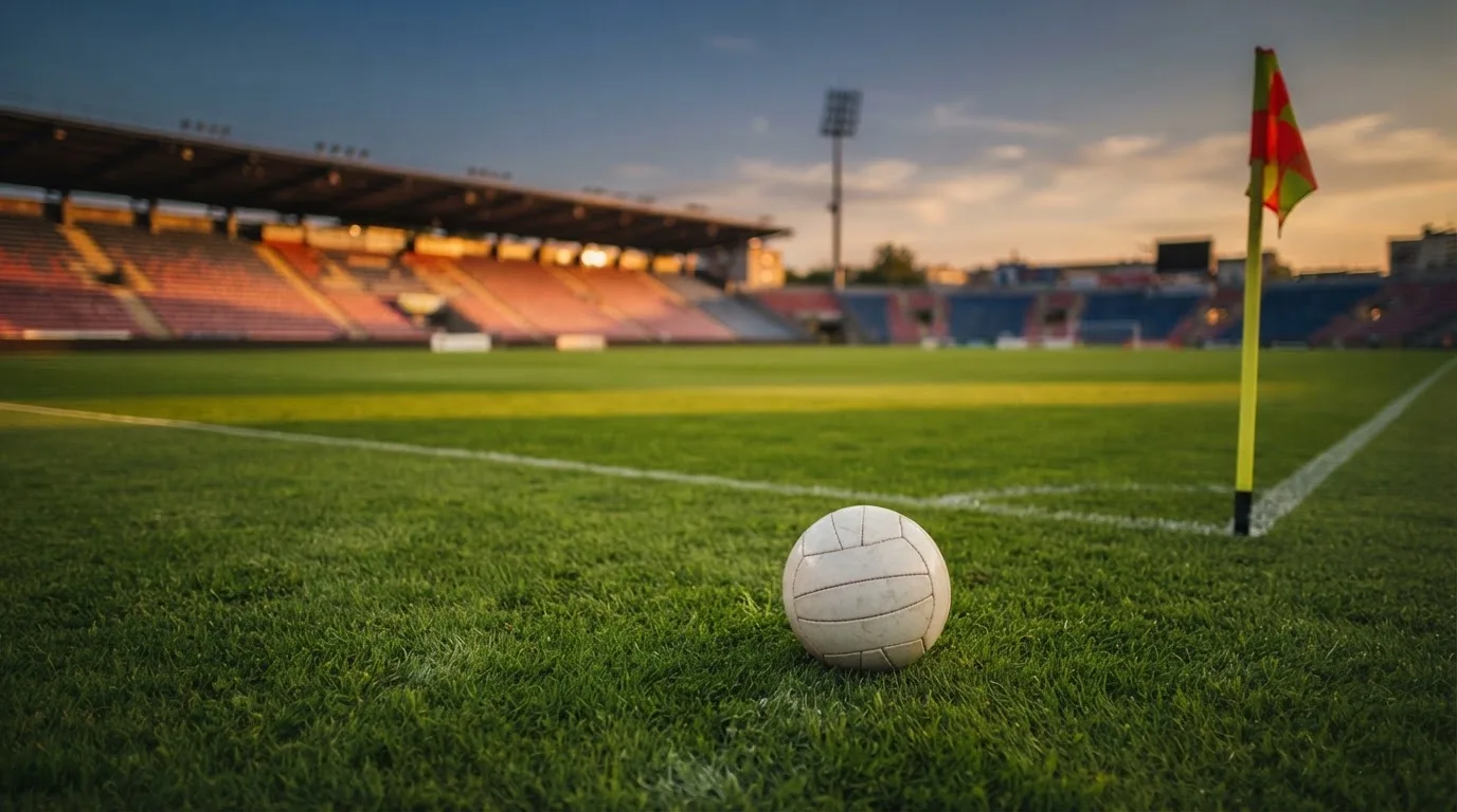 Ein Fußball liegt auf dem Rasen neben der Eckfahne eines leeren Platzes, dahinter eine Tribüne im Abendlicht.