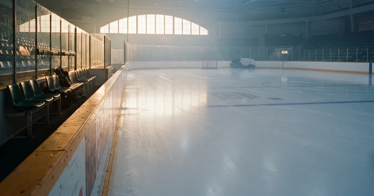 Eine zugefrorene Eisfläche am Rand eines Eishockeyfelds im Morgenlicht, Banden und leere Spielerbänke von der Seite fotografiert.