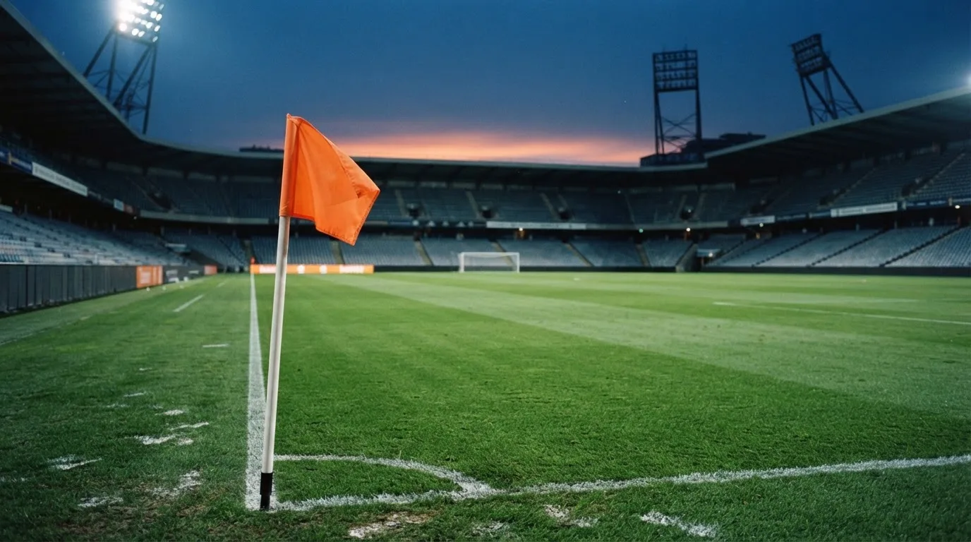 Eine Eckfahne am Rand eines Rasenplatzes, dahinter ein leeres Stadion unter Flutlicht aus der Ferne gesehen.