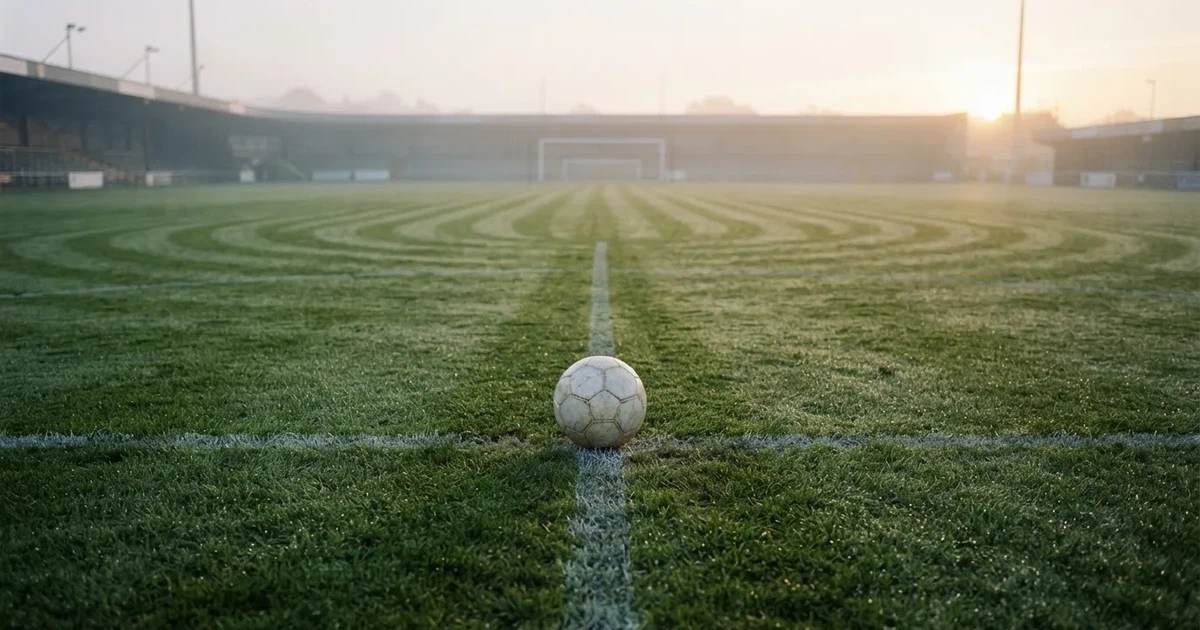 Ein Fußball liegt auf dem Anstoßpunkt eines leeren Rasenplatzes in der Morgendämmerung, Tau glitzert auf dem Gras.