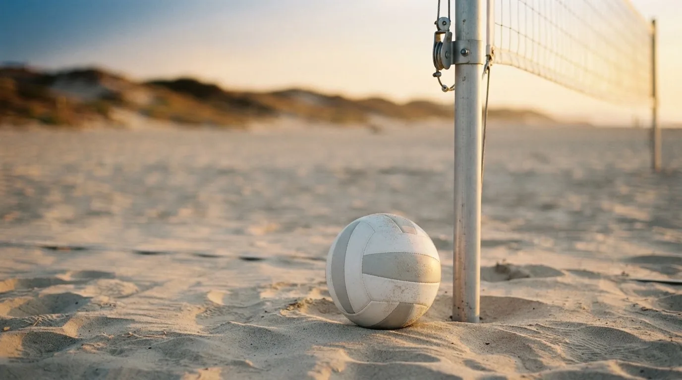 Ein Volleyball liegt auf Sand neben einem Netzpfosten, Strandlicht am späten Nachmittag, im Hintergrund unscharfe Dünen.