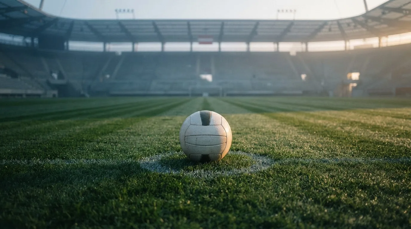 Ein Fußball auf dem Mittelpunkt eines Rasenplatzes, früher Morgen, Tau auf dem Gras, im Hintergrund leere Tribünen.