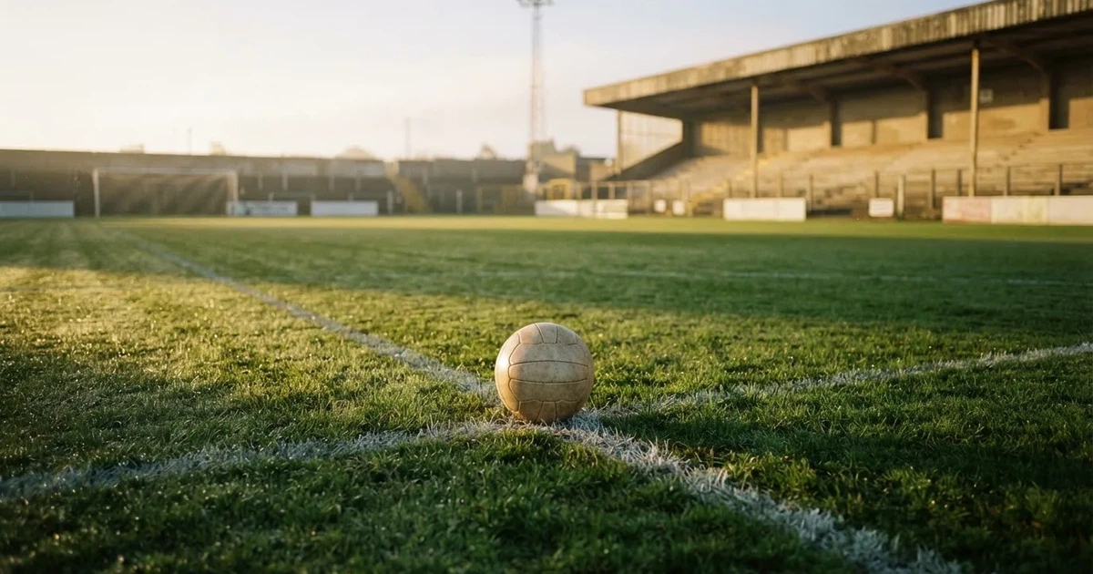 Ein Fußball liegt auf dem Elfmeterpunkt eines leeren Rasenplatzes im Morgenlicht, dahinter eine ruhige Tribüne ohne Zuschauer.