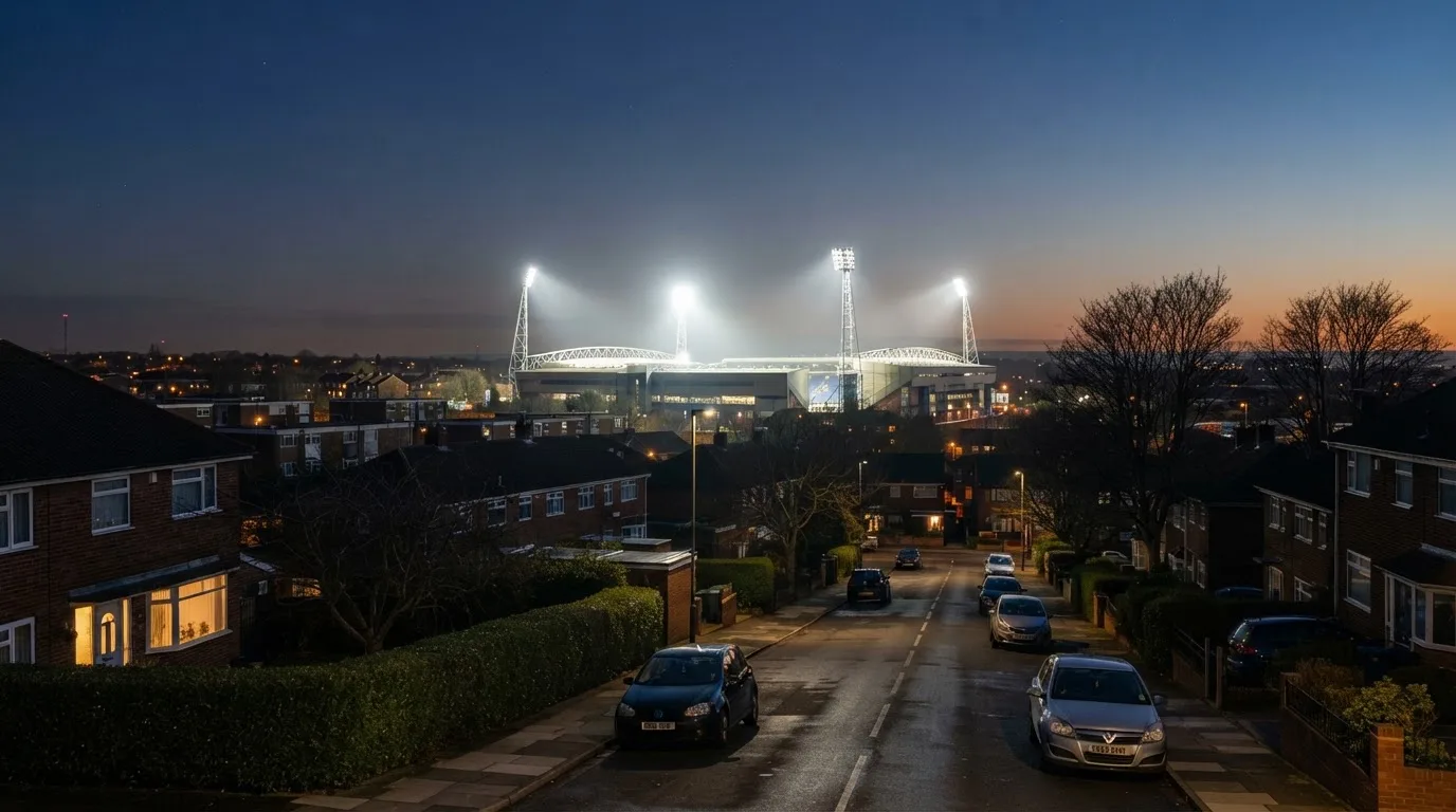 Ein Flutlichtstadion bei Dämmerung aus weiter Ferne gesehen, die Lichtmasten leuchten über Dächern, Bäumen und einer ruhigen Stadtrandlandschaft.