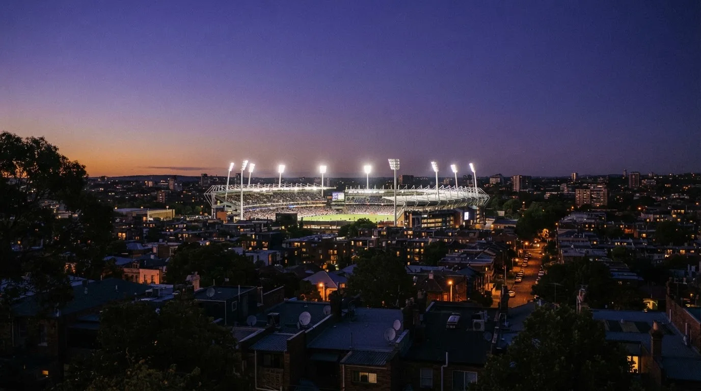 Ein Flutlichtstadion bei Dämmerung aus weiter Ferne gesehen, die Lichtmasten leuchten über Dächern und Bäumen.