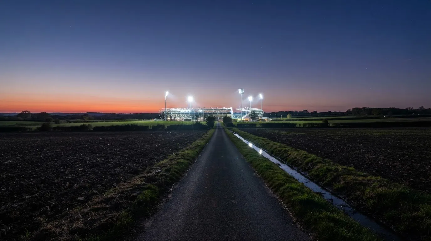 Ein Stadion unter Flutlicht aus der Ferne gesehen, davor eine dunkle Landschaft mit Feldern und einer Landstraße.