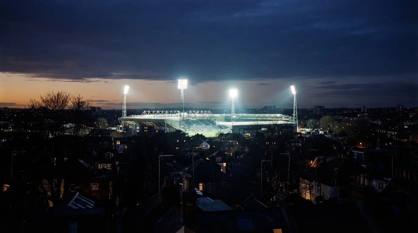 Ein Flutlichtstadion bei Dämmerung aus weiter Ferne gesehen, die Lichtmasten leuchten über Dächern und Bäumen.